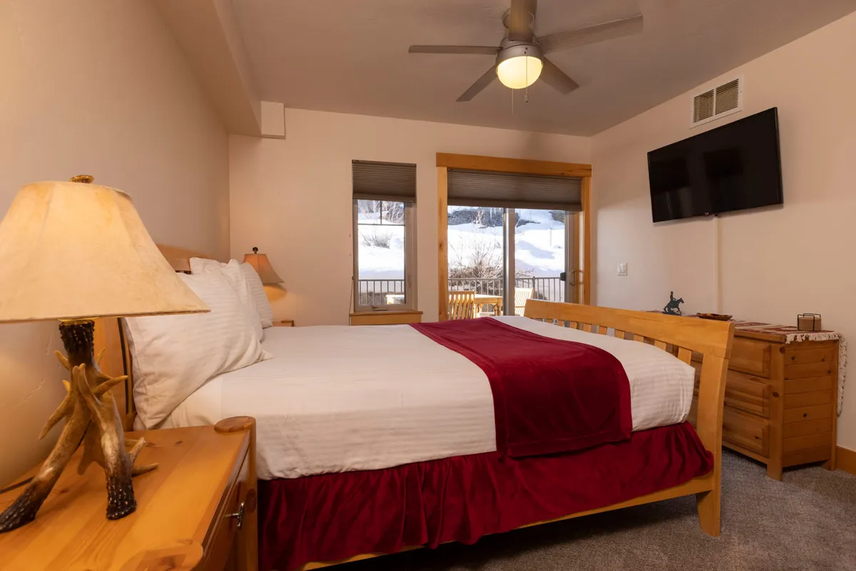A spacious bedroom with a plush bed adorned with a red throw, warm lighting from a bedside lamp, and large windows that offer a scenic view outdoors. The room is styled for comfort and relaxation.