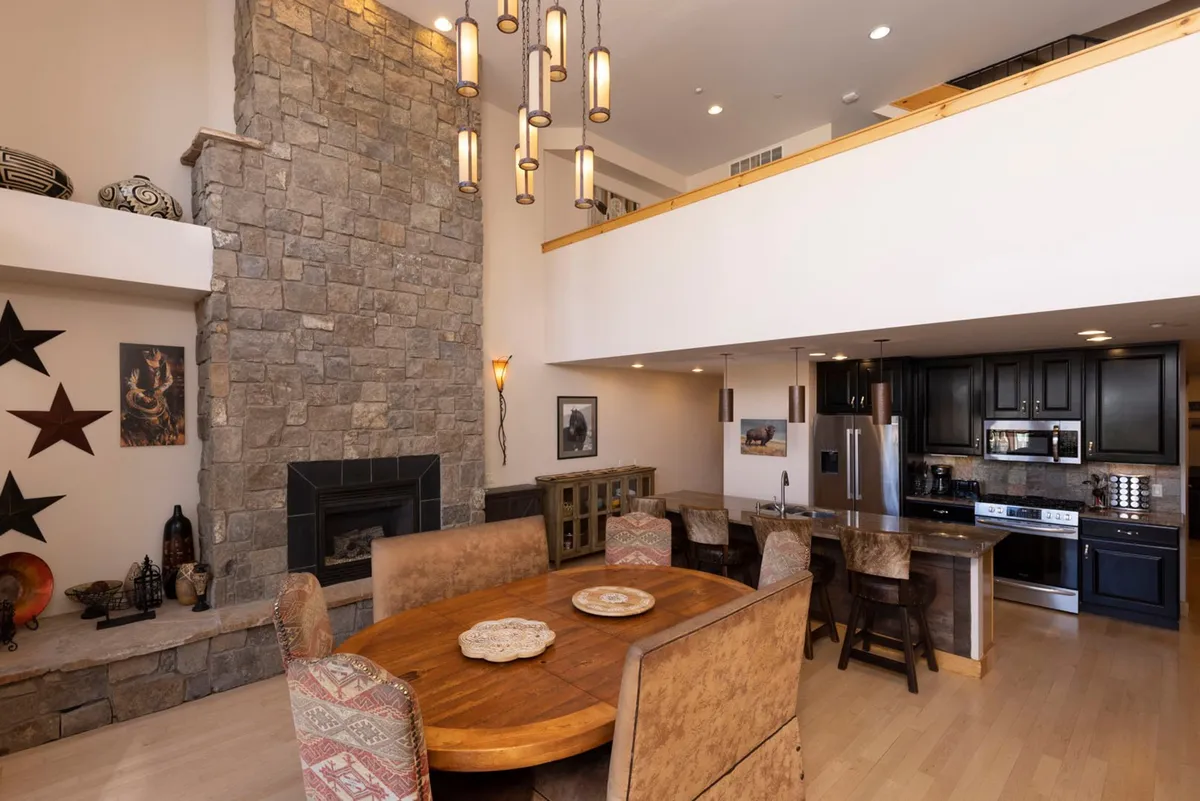 An open-plan space featuring a beautiful stone fireplace, a dining table set for group meals, and a glimpse of the adjoining kitchen area. The ambiance is inviting and warm, ideal for socializing and celebrating.