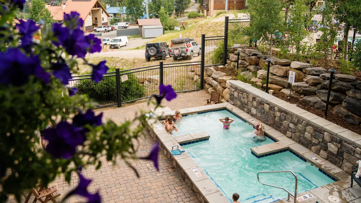 A steaming hot tub and sparkling swimming pool sit outside, bordered by stone walls and lush flowers. The inviting space offers an ideal setting for relaxation or socializing after a day on the slopes, with mountain air and nature all around.