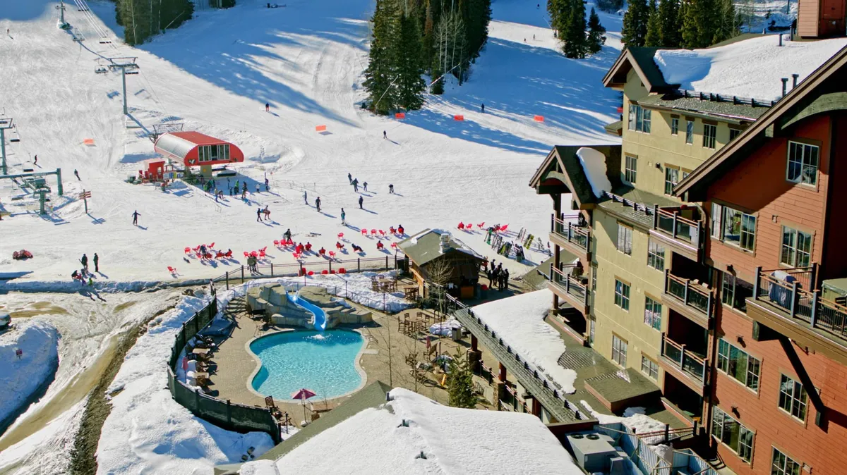 A lively winter scene featuring snow-covered ski slopes directly adjacent to the resort's lodge buildings, with a ski lift and skiers in the foreground. The outdoor pool adds a family-friendly winter resort atmosphere, and the expansive snowy landscape emphasizes direct slope access.
