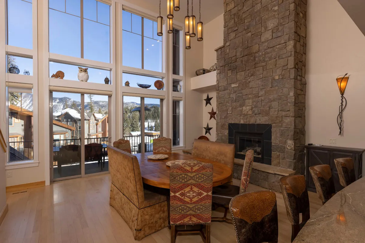 The image showcases the inviting interior of the townhome, centered around a glowing stone fireplace, expansive windows framing mountain views, and a spacious dining table set for gatherings. The scene evokes warmth and togetherness, perfect for family meals or relaxing après-ski moments.
