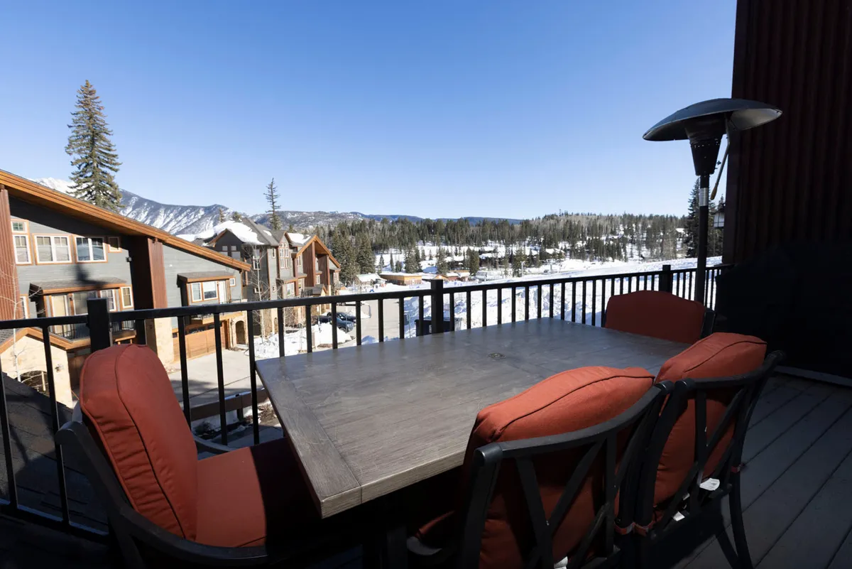 An outdoor dining table sits on a snow-dusted patio, overlooking a breathtaking mountain view. The area is surrounded by a pristine winter landscape, with slopes visible in the distance, evoking a sense of peaceful retreat and direct connection to the outdoors.