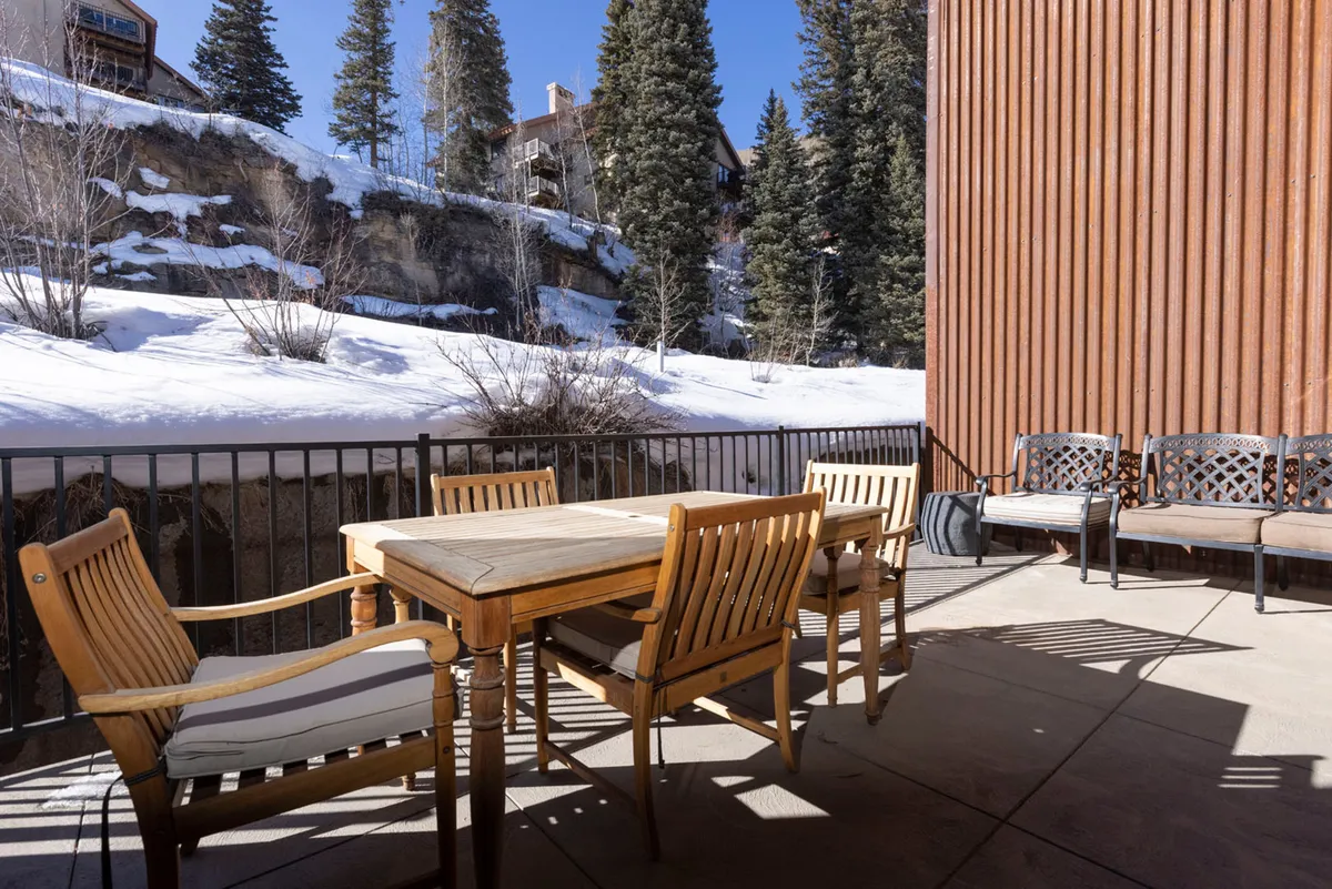 A rustic wooden dining table and chairs set outdoors, surrounded by a snowy landscape and tall pine trees. The scene conveys a sense of camaraderie and the potential for shared meals or drinks with a view of the action on the slopes.