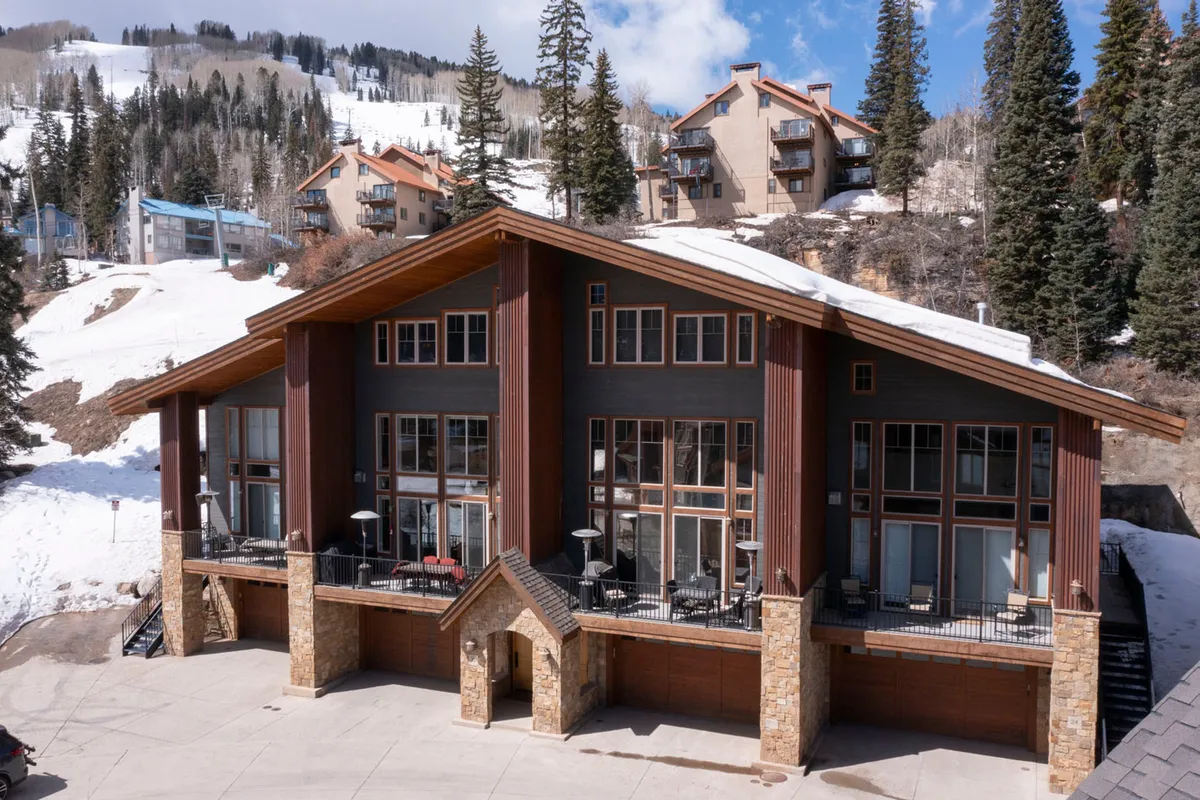 A welcoming view of the lodge's exterior featuring large windows, a stone facade, wooden accents, and a snow-covered landscape. The cozy atmosphere is evident, with the building nestled into the scenery, suggesting a perfect family retreat after outdoor fun.