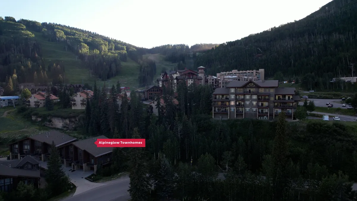 A wide exterior shot featuring the Alpenglow Townhomes set against a backdrop of snow-covered mountains and tree-lined slopes. The surrounding resort buildings hint at convenient access to skiing and group adventures, while the tranquil lighting suggests a welcoming environment, perfect for gathering after a day on the slopes.