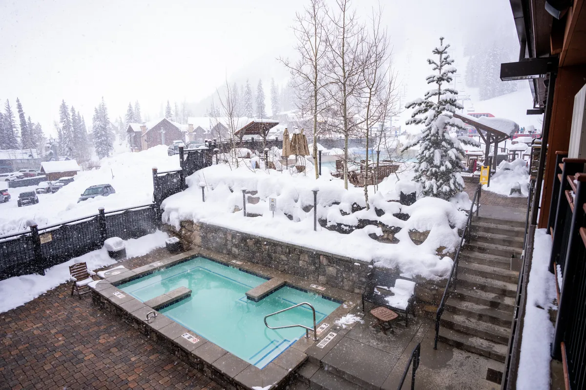 An inviting outdoor hot tub set amidst a snow-covered landscape and framed by a rustic stone wall. Snow-laden trees in the background create a cozy winter ambiance, suggesting a perfect spot for groups to relax and socialize after a day on the slopes.