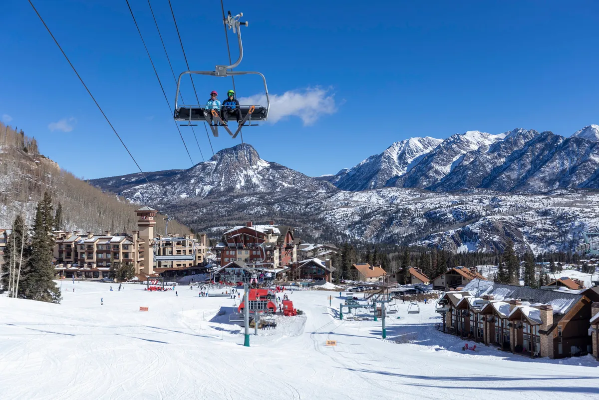 A panoramic view of the ski lift, snow-covered slopes bustling with skiers and snowboarders, with a majestic mountain backdrop and resort buildings. The image conveys the excitement and accessibility of winter sports right outside the accommodation.
